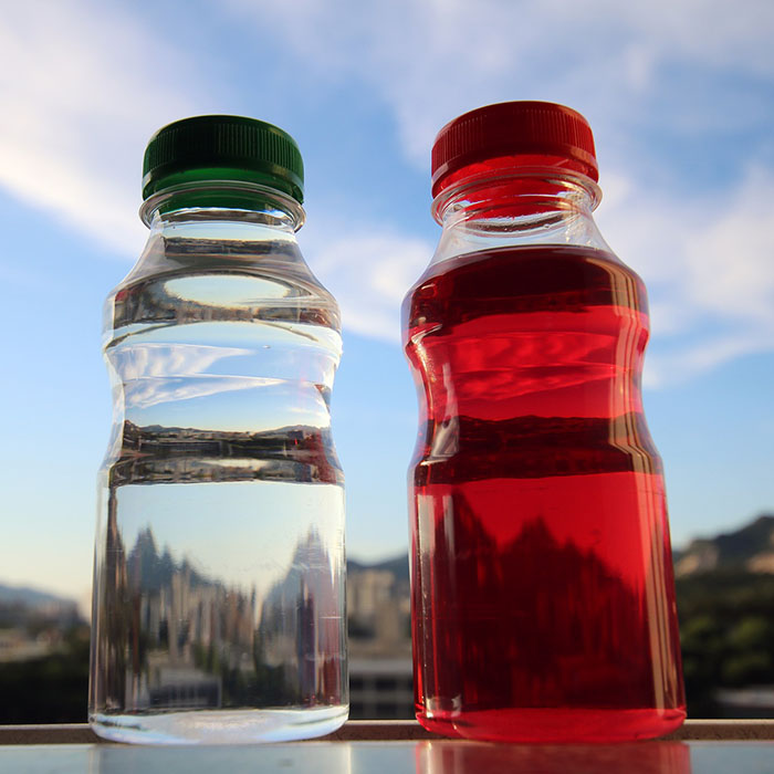 Oral Health and What We Drink, closeup of 2 clear bottles with the blue sky as a backdrop, the bottle on the left has water, the bottle on the right contains a red liquid like a sports drink
